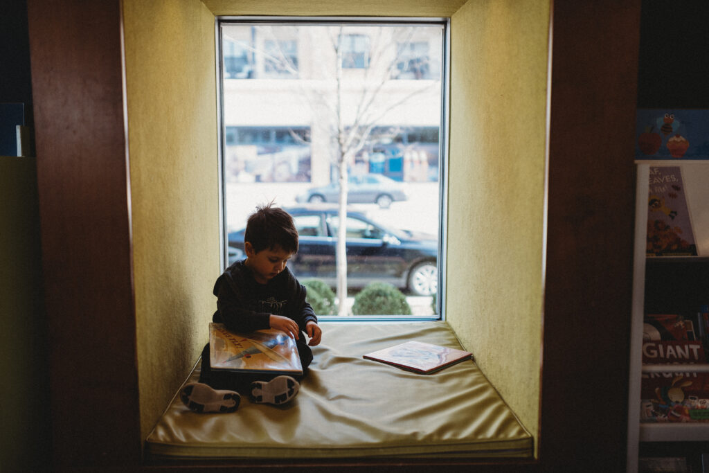 Toddler boy sits in the library window reading books, as an example of family photo location ideas. 